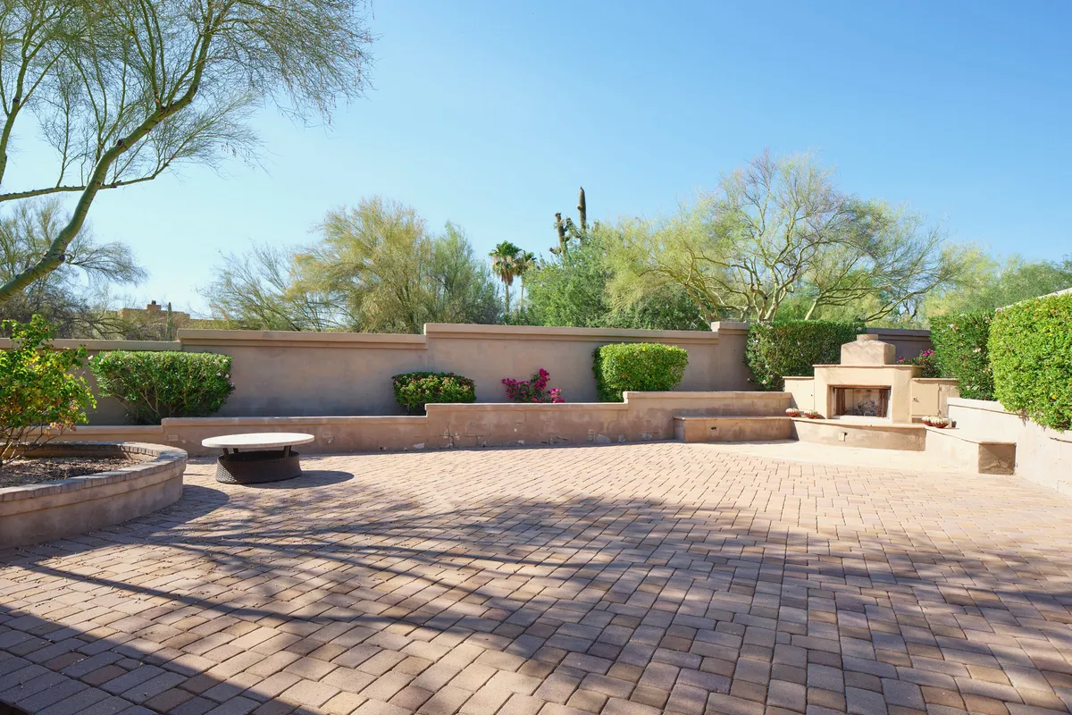 A tranquil exterior patio scene featuring a modern outdoor fireplace, surrounded by lush greenery and paved stonework. The setup is ideal for evening gatherings, stargazing, or enjoying the warmth and ambiance of the fire after a day in the sun.