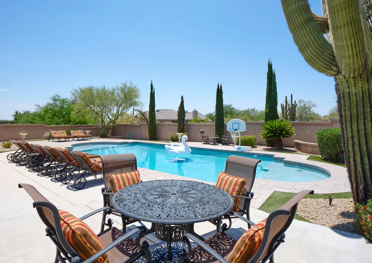 This image features an inviting backyard with a sparkling swimming pool, comfortable outdoor seating, a decorative cactus, and a basketball hoop. The lush green lawn and landscaping create a welcoming space ideal for both relaxation and play.