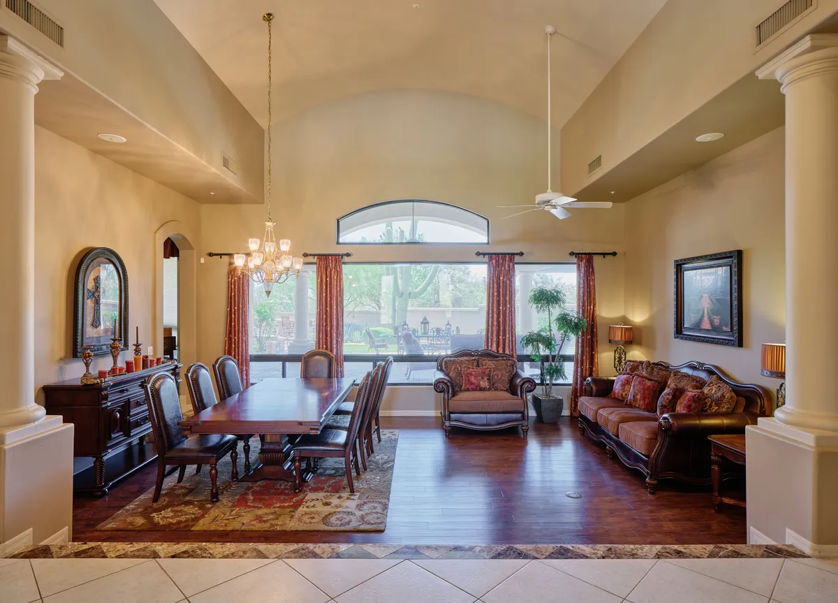 A spacious, open-plan dining and living area featuring a large dining table that comfortably seats several guests, a sofa area for lounging, a chandelier providing elegant lighting, and expansive windows that let in natural light. The layout emphasizes the communal aspect of meals and gatherings.