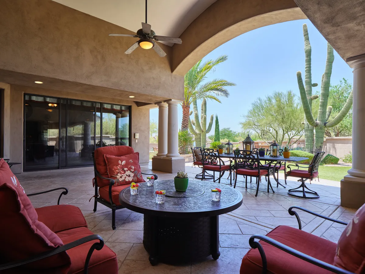 A patio set beneath a shaded area, surrounded by desert-friendly plants and cacti. There is a comfortable seating area and an outdoor dining table, set against a tranquil, sunlit backdrop.