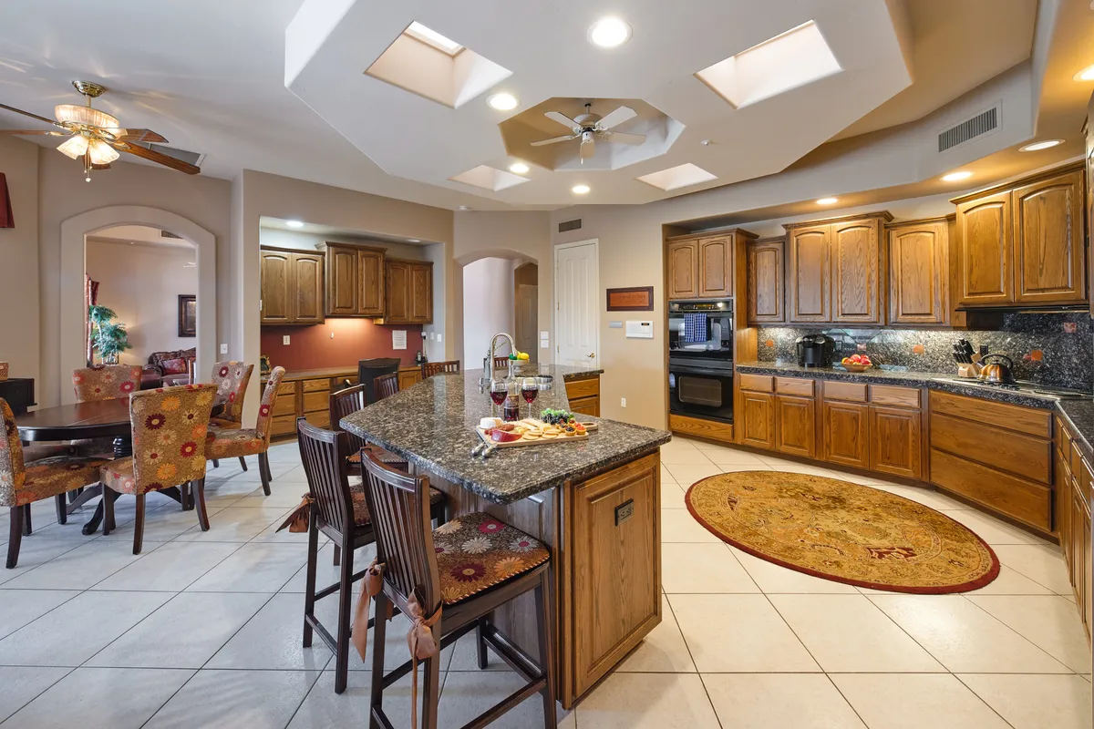 This image features a spacious kitchen with a prominent granite island, colorful dining chairs around a table, rich wood cabinetry, and skylights that bathe the room in natural light. The island provides bar seating and serves as a central gathering spot for cooking and casual conversation.