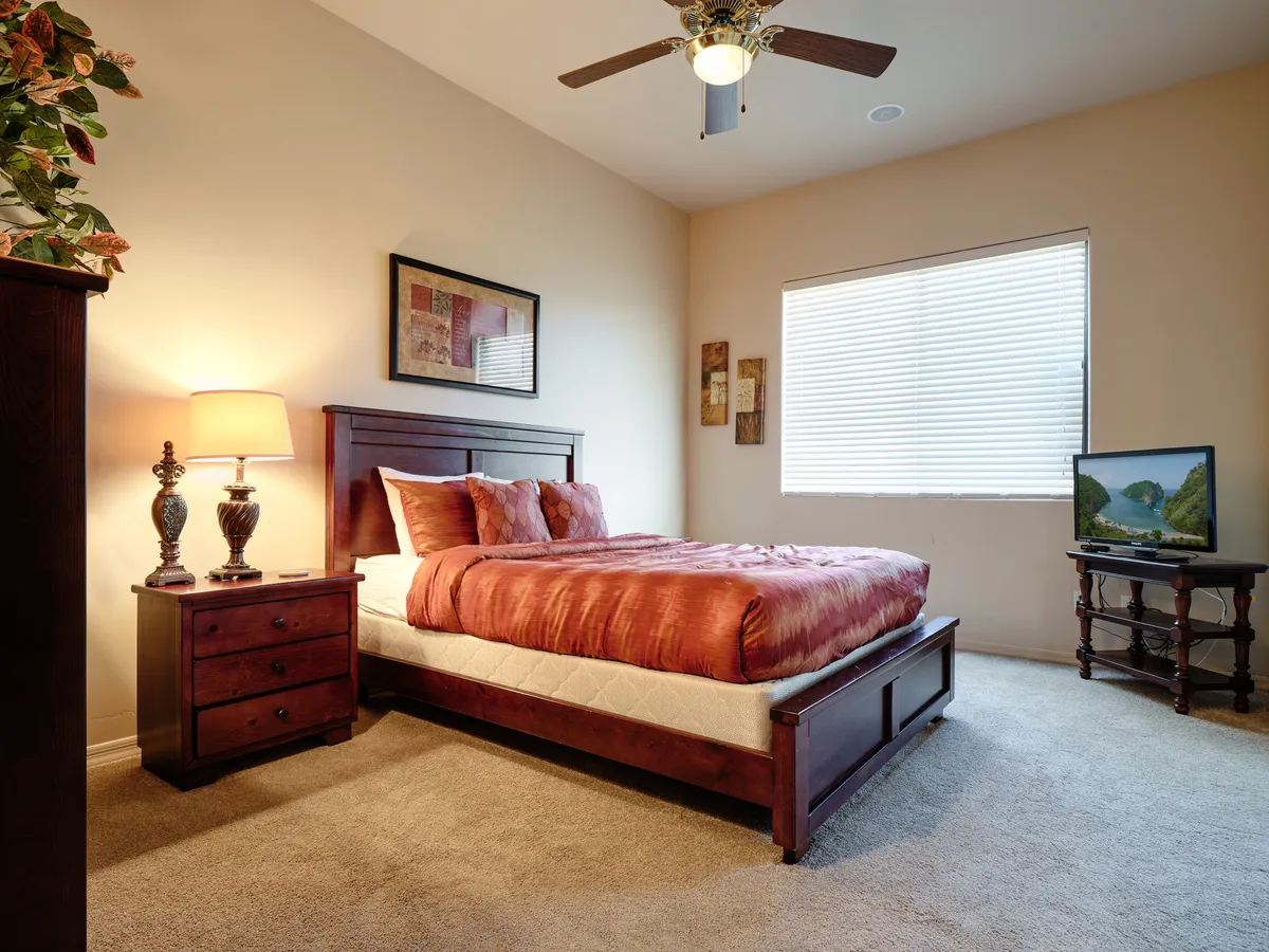 A guest bedroom with a bed adorned with decorative pillows, a nightstand with a lamp, and a TV on a stand facing the bed. Natural light streams in through the window with blinds, emphasizing a cozy and relaxing environment.