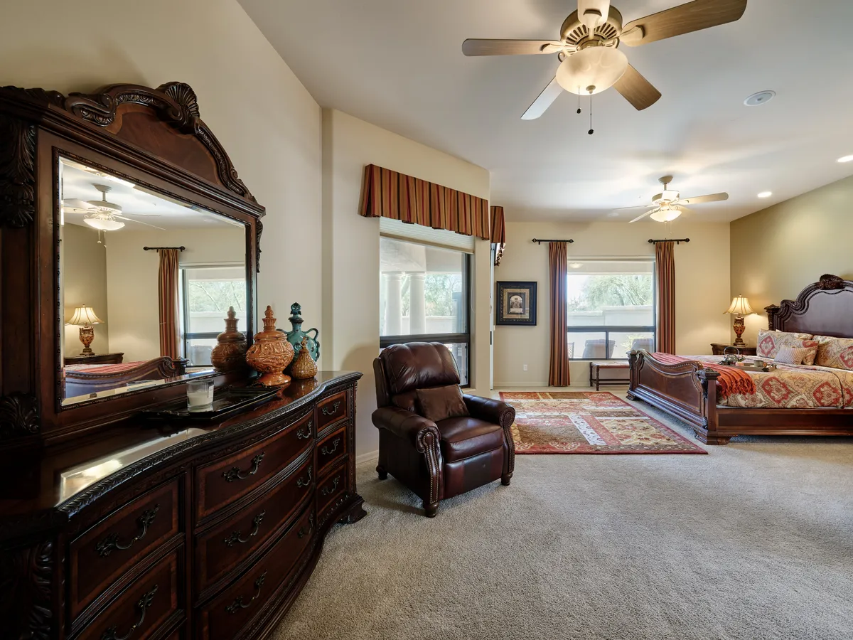 A tranquil bedroom featuring an ornate wooden dresser with a mirror, a comfortable leather chair, and a decorative bed with patterned bedding. A window allows natural light to fill the space, and the atmosphere is serene and inviting.