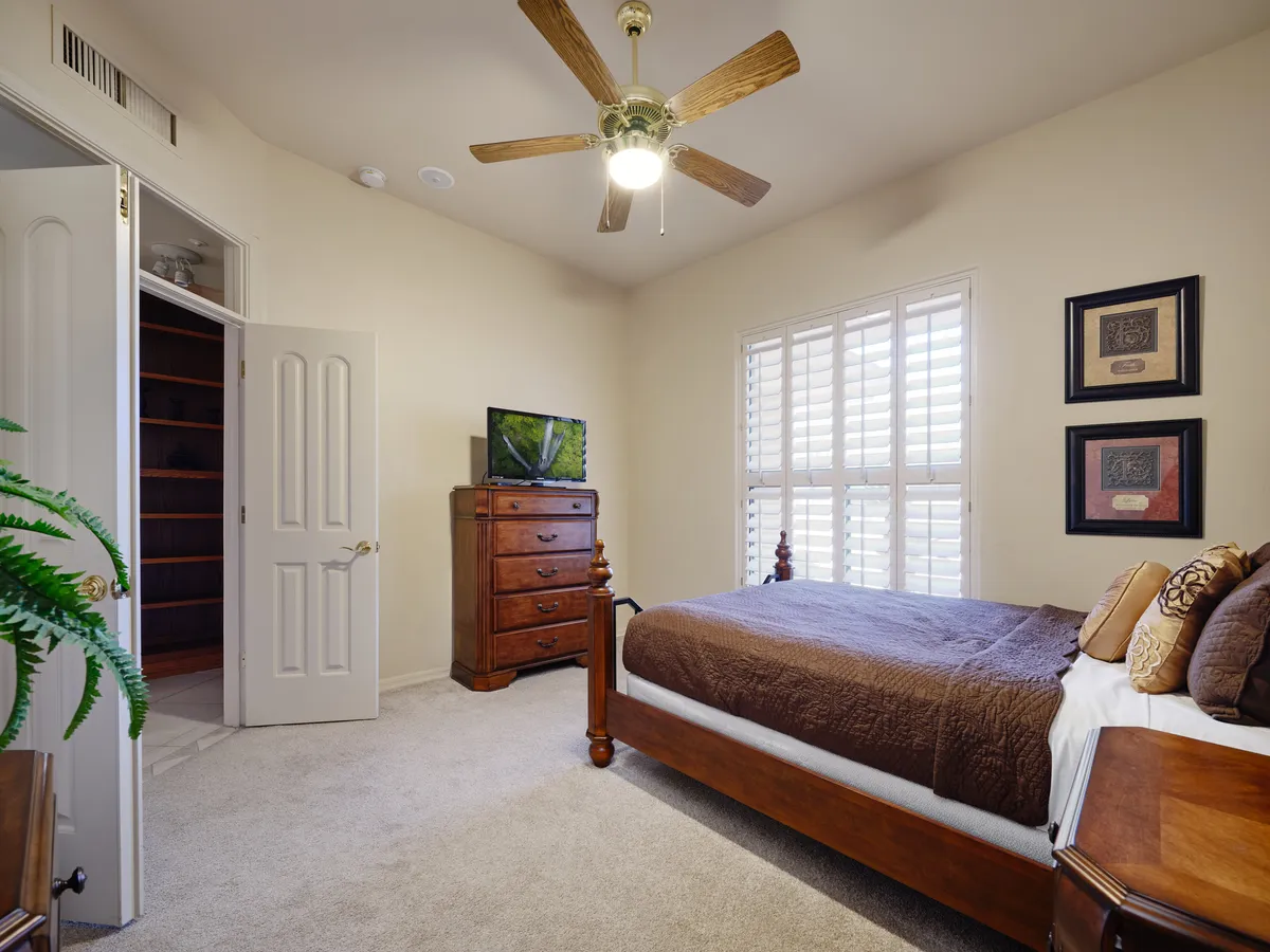 A serene bedroom setup with a bed dressed in brown bedding, a chest of drawers with plants, a television, and a window with shutters letting in sunlight. The space feels peaceful and private, ideal for quiet moments or watching TV in bed.