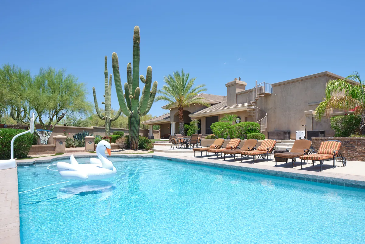 The hero image showcases the backyard of Valley View Villa at sunset, featuring a sparkling swimming pool surrounded by lounge chairs, playful cacti and palm trees, and an eye-catching inflatable swan floating on the water. The scene captures the relaxed ambiance of evening gatherings, with hints of children playing and adults enjoying the desert landscape.