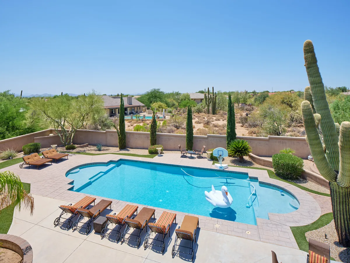 A tranquil backyard scene featuring a sparkling swimming pool with a swan float, lounge chairs, desert landscaping, and plenty of space for outdoor gatherings. The setting invites sunbathing, swimming, and relaxed entertaining under the desert sky.