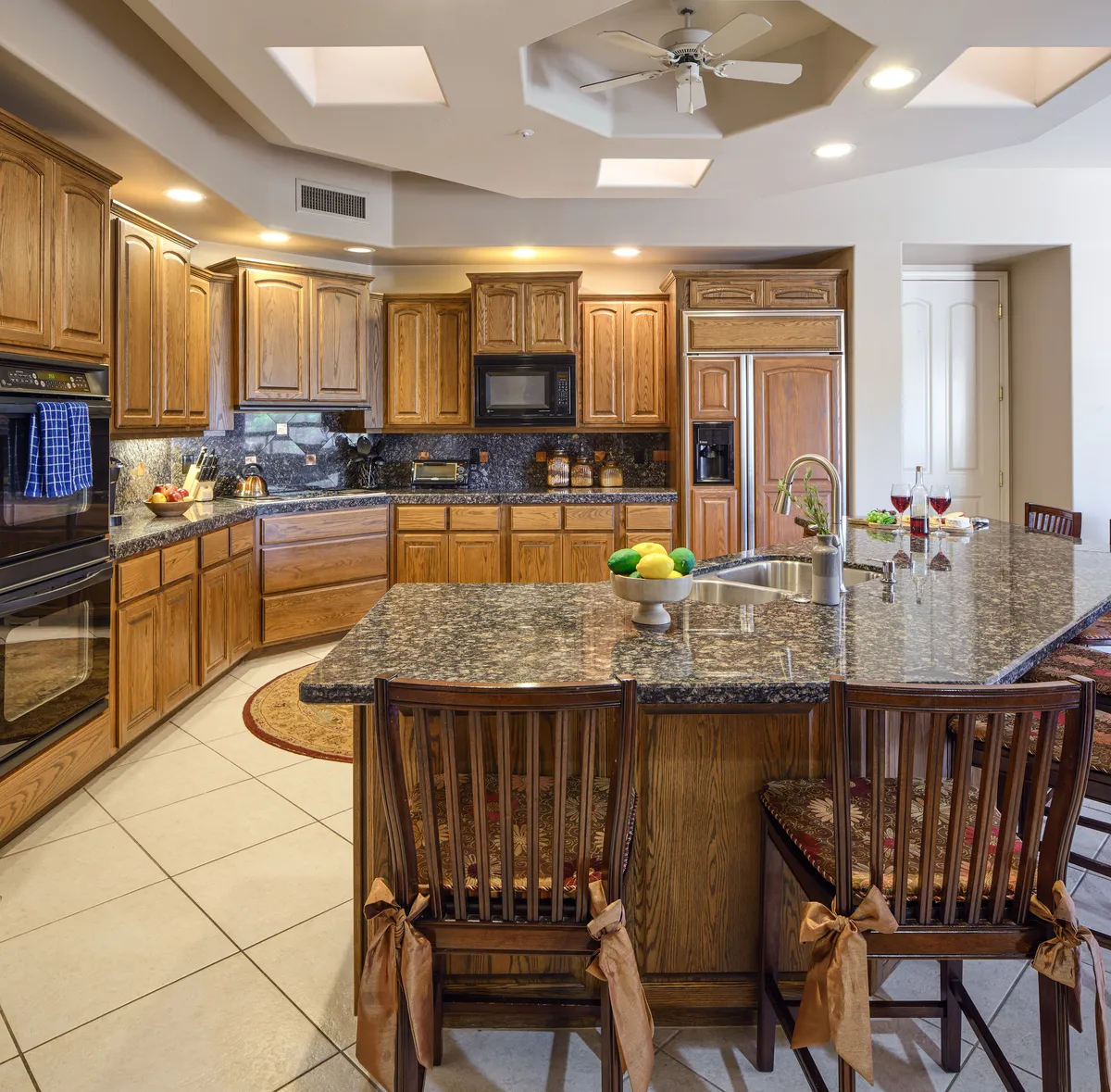 A warm, modern kitchen space showcasing a granite countertop, wooden cabinets, a central kitchen island with bar stools, and ample space for meal prep and gathering. The design is welcoming, ideal for both cooking and socializing.
