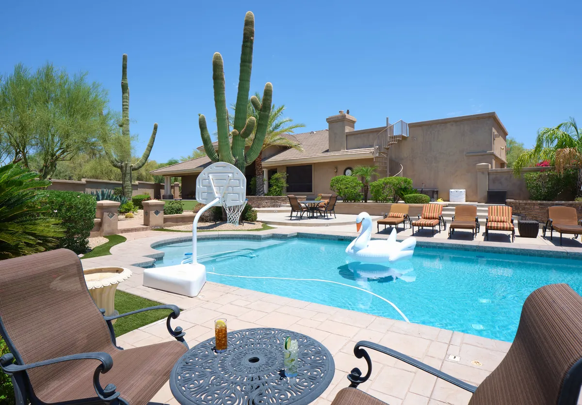 This exterior image showcases a sparkling swimming pool equipped with a basketball hoop, surrounded by lawn furniture and a saguaro cactus. A large swan float adds a playful touch, while the setting sun casts a warm glow over the inviting scene. The space is clearly designed for leisure, games, and gatherings.